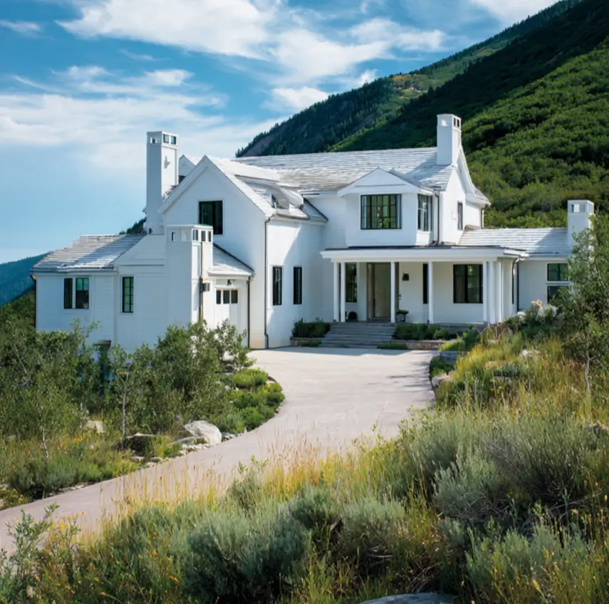 A large white modern mountain house with black window trim, set on a sprawling green hillside under a bright blue sky.