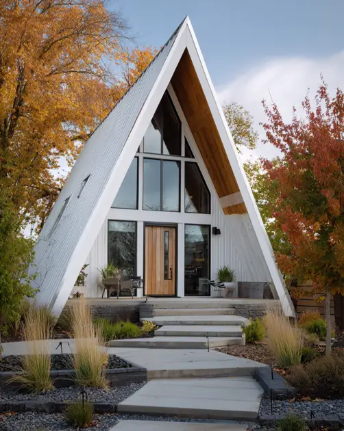 Tiny A-Frame Cabins: 31 Inspiring Ideas for Your Dream Retreat 8 A striking small a-frame house with a geometric white exterior and a full glass front surrounded by autumn trees.