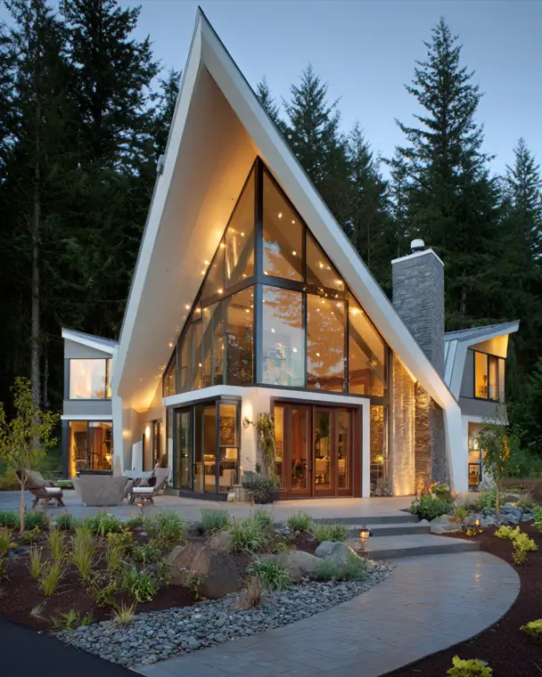 A contemporary A-frame house with a soaring white roofline, glass facade, and stone accents, illuminated against a forest at dusk.