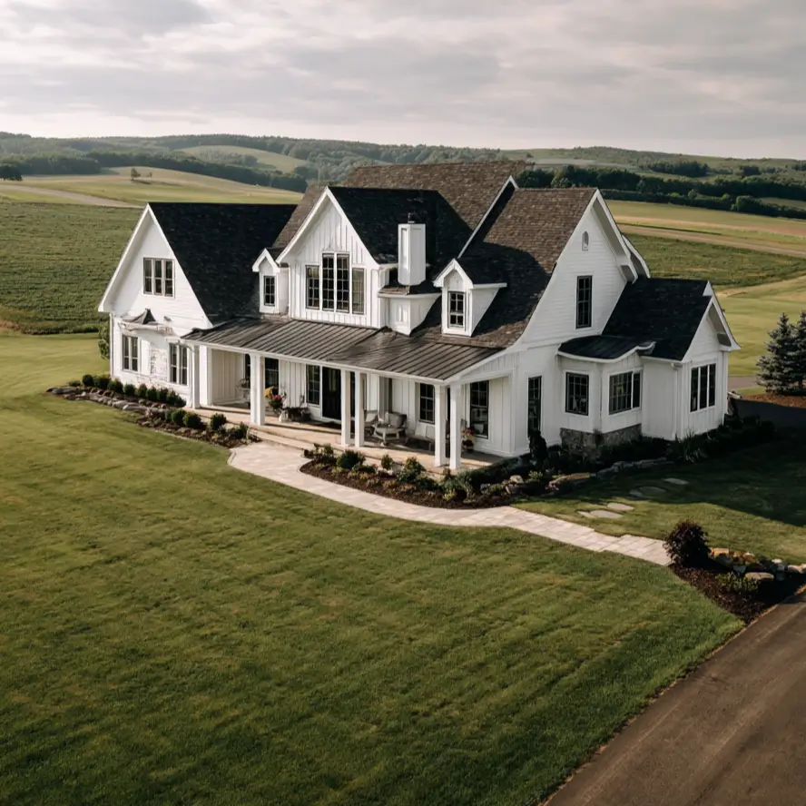 An aerial view of a grand white farmhouse with a wrap-around porch, nestled in a landscape of rolling green hills.