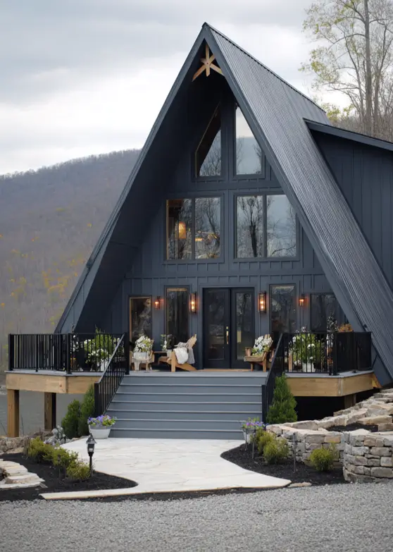 A dark charcoal modern a frame cabin featuring a large elevated deck with outdoor seating overlooking a hilly landscape.