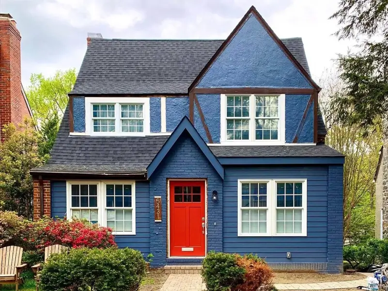 Striking small modern Tudor-style house with a bold, monochromatic deep blue exterior, crisp white window trim, and a high-contrast red front door.