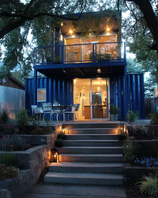 A two-story blue shipping container home illuminated at dusk, nestled under trees, with lighted stone steps leading to a ground-floor patio and an upper balcony.