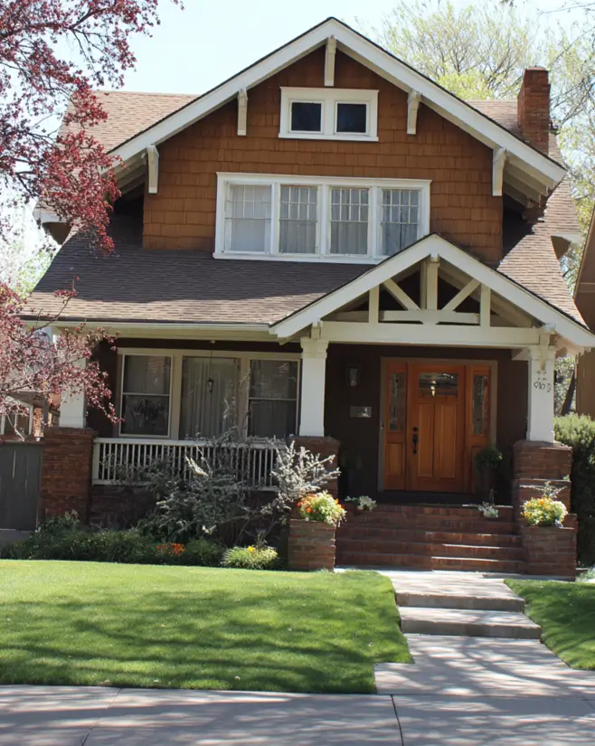 A classic Craftsman suburban house exterior with warm brown shingle siding, a deep front porch supported by white tapered columns set on sturdy brick piers.