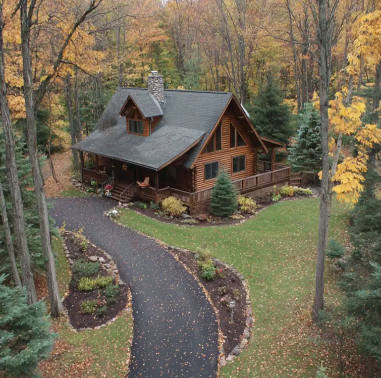A log cabin with a wraparound porch, nestled in a vibrant autumn forest with a curving driveway.