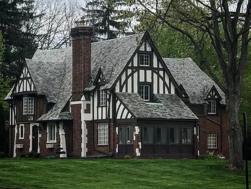 A classic Tudor style house exterior featuring a prominent brick chimney, dark half-timbering, and a gray slate roof, set on a green lawn.