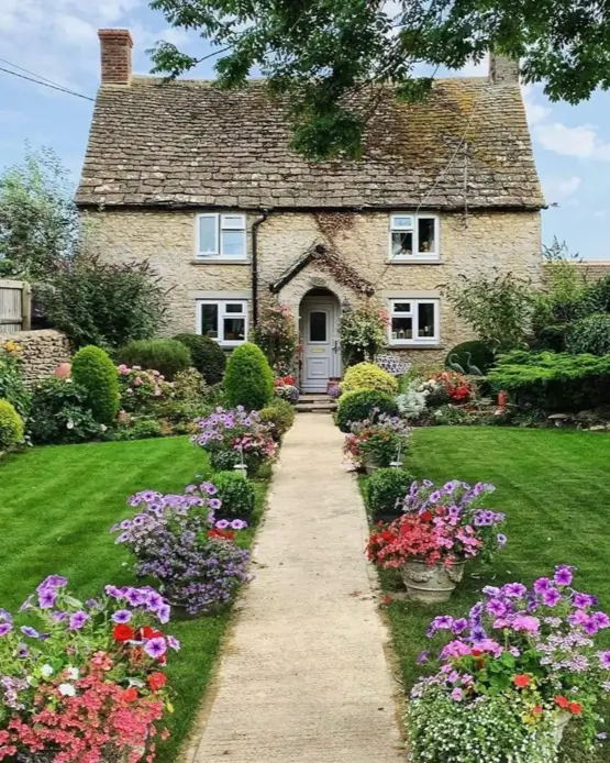Quintessential english cottage house design featuring a symmetrical stone path bordered by vibrant purple and red flowers leading to a classic Cotswold stone home.