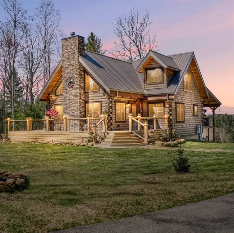 A stunning log cabin home design at sunset, featuring a large fieldstone chimney, a wraparound deck, and a metal roof.