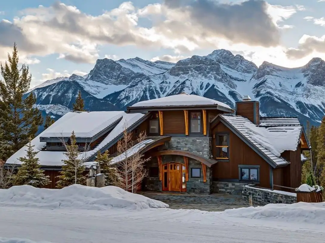 A luxury modern mountain house with a wood and stone exterior, covered in snow, set against a dramatic snow-capped mountain range.