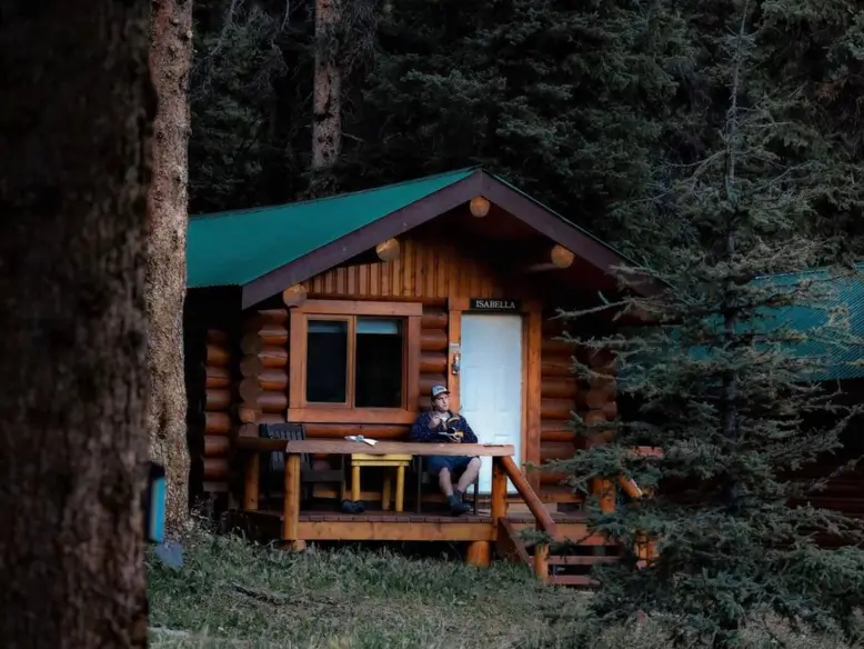 A man relaxes on the porch of a small, rustic tiny log cabin, surrounded by a dense and peaceful pine forest.