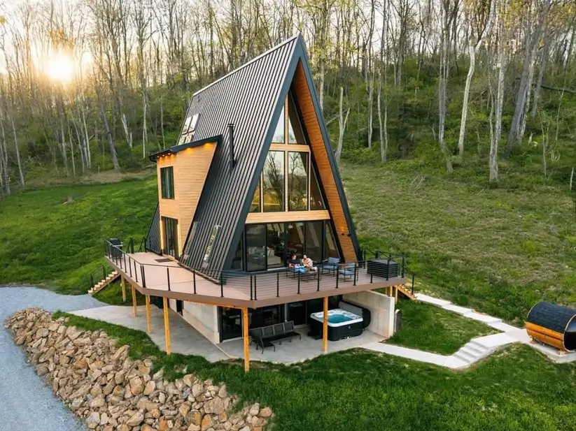A modern a frame home with a black metal roof and wood siding, featuring a large wraparound deck with a hot tub, set on a sunny hillside.