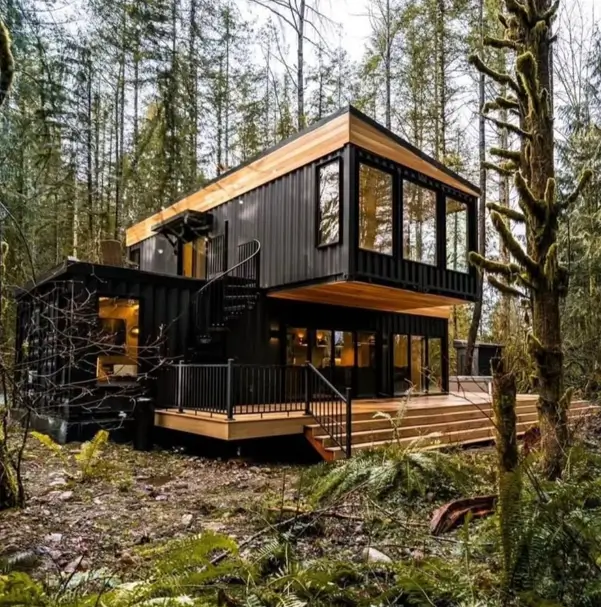 A modern two-story black shipping container home with warm wood accents, featuring an external spiral staircase and a large deck, nestled in a lush, mossy forest.