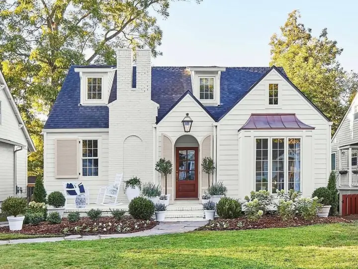 A modern cottage house exterior with creamy white siding, a prominent brick chimney, a dark blue roof, and an elegant copper awning over the bay window.