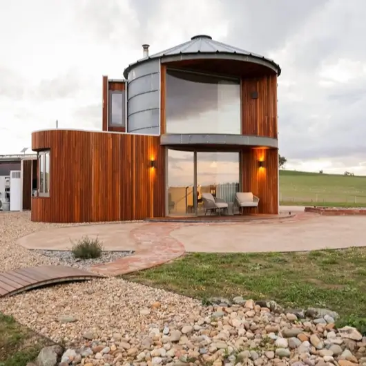 A stunning modern grain silo home featuring a mix of warm wood cladding and metal, with large windows overlooking a landscaped patio at dusk.