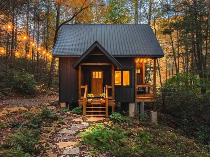 An inspiring tiny cabin idea featuring a modern black exterior and a metal roof, with warm string lights creating a cozy ambiance in a forest at dusk.