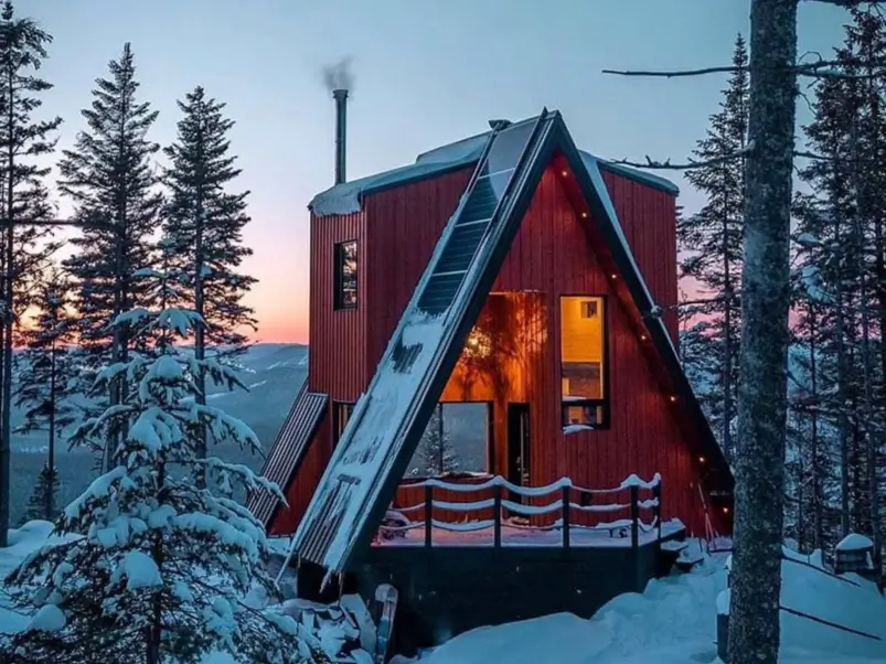 A modern red tiny a frame cabin covered in snow at dusk, with solar panels on the roof and warm lights glowing from inside a winter forest.