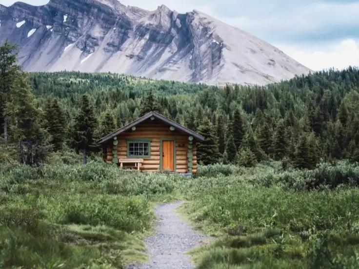 A small, secluded tiny log cabin sits in a lush green meadow at the base of a large, majestic mountain, representing an escape to nature.