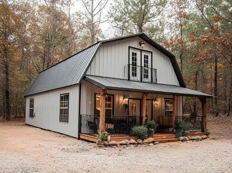 A small barndominium home with white siding and a gambrel roof, featuring a wooden porch and a small balcony in a wooded setting.