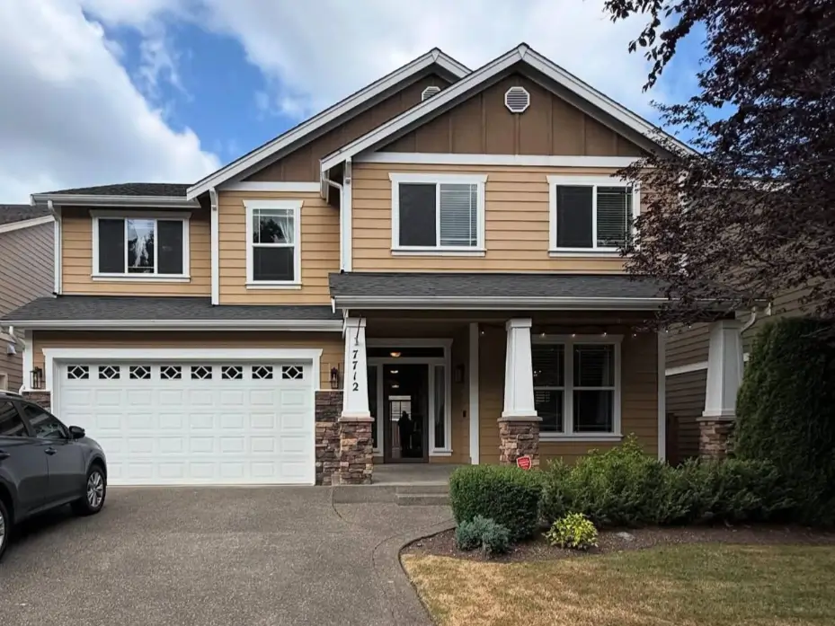 A classic suburban house exterior featuring warm tan siding, a covered front porch with stone-based columns, and a white garage door, creating a welcoming and timeless curb appeal.