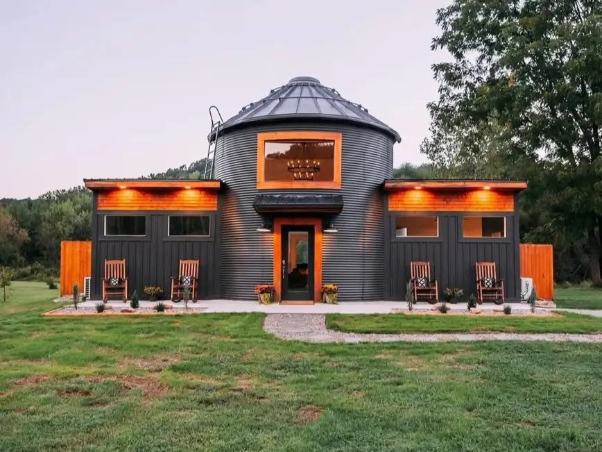 A modern grain silo house with a dark corrugated metal finish, serving as the central point between two symmetrical modern extensions.