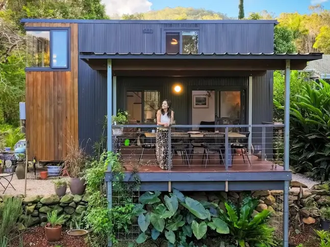 A woman enjoying her modern tiny house with a covered deck surrounded by lush greenery.