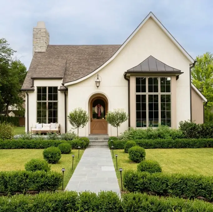 A modern beige house with black trim, featuring an arched wooden door and manicured landscaping.