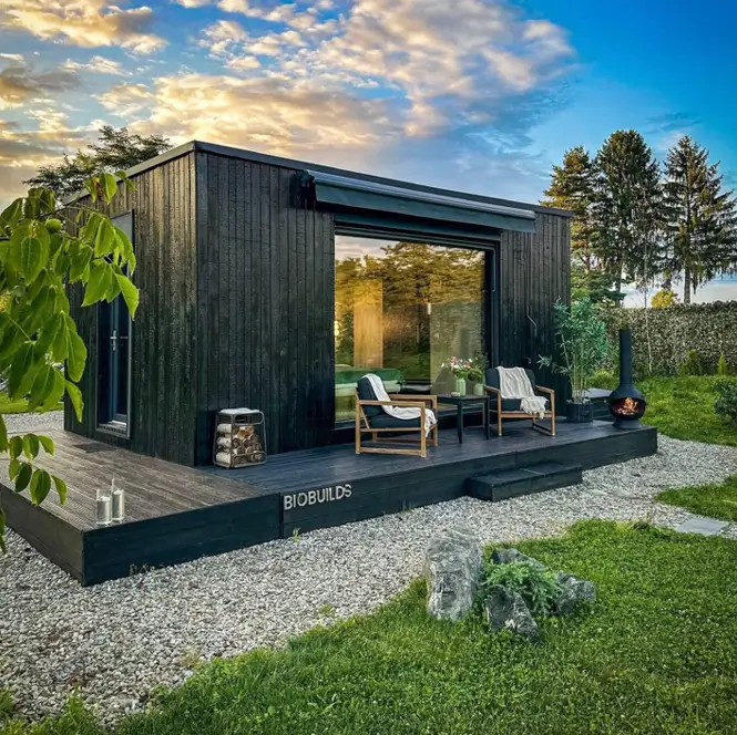 Modern black minimalist cabin with a large window and a dark wood deck in a garden setting at sunset.