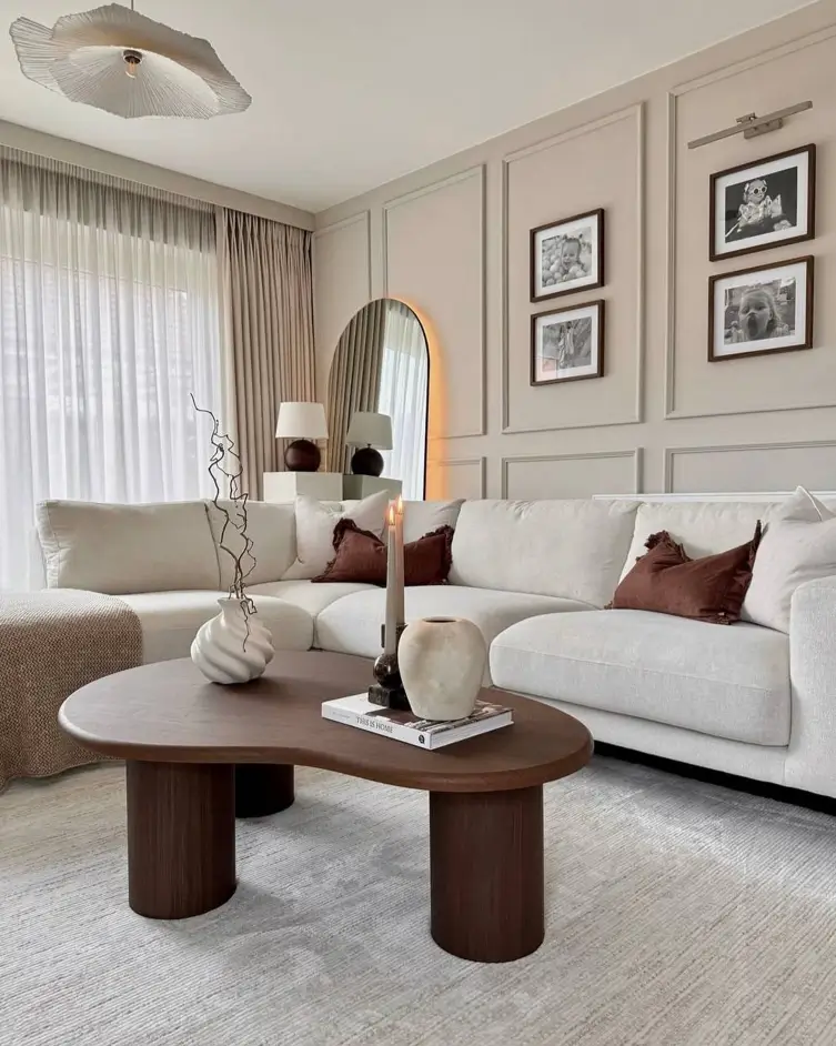 A warm neutral living room with an off-white sectional, a dark wood coffee table, rust-colored pillows, and an arched mirror.