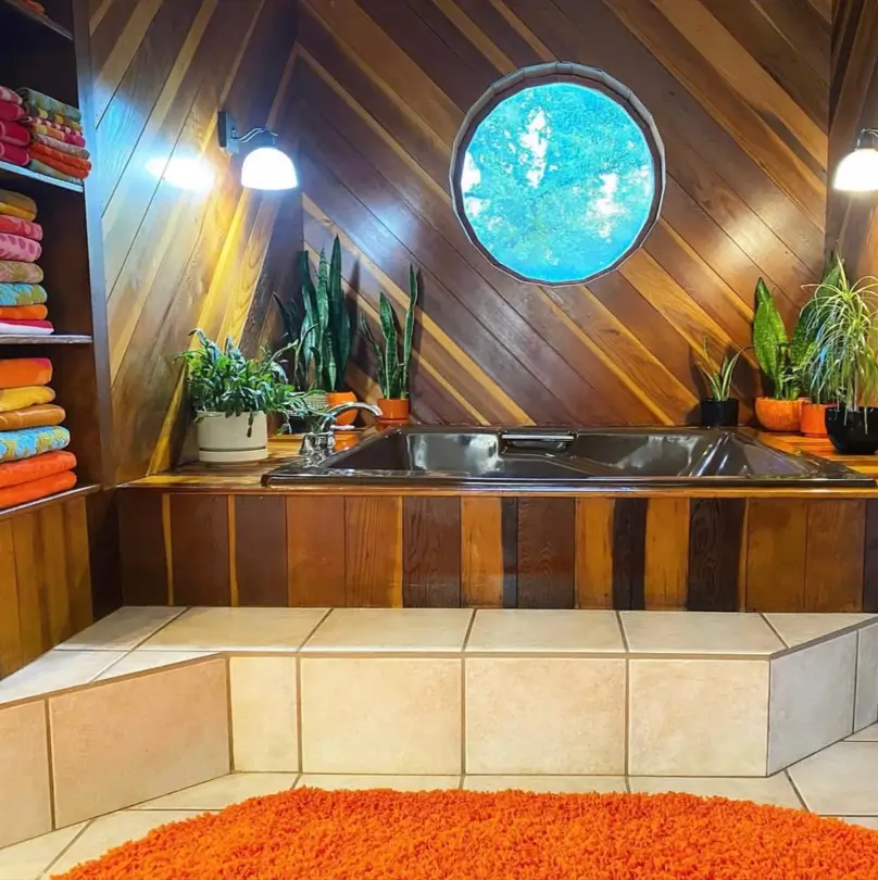 A 70s bathroom idea with diagonal wood paneling, a round porthole window, and an orange shag rug next to a sunken tub.