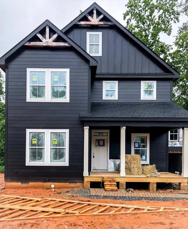 A black farmhouse under construction with white trim and decorative light-wood trusses in the gables.
