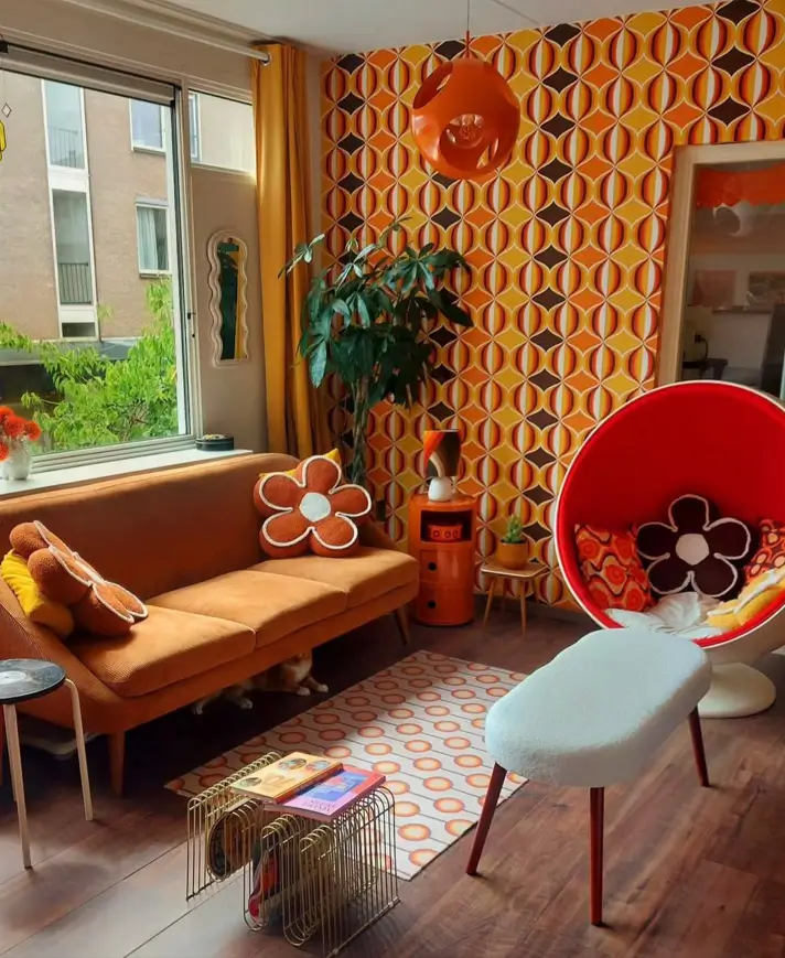 A 70s living room with bold geometric wallpaper, an orange sofa, and a red pod chair.