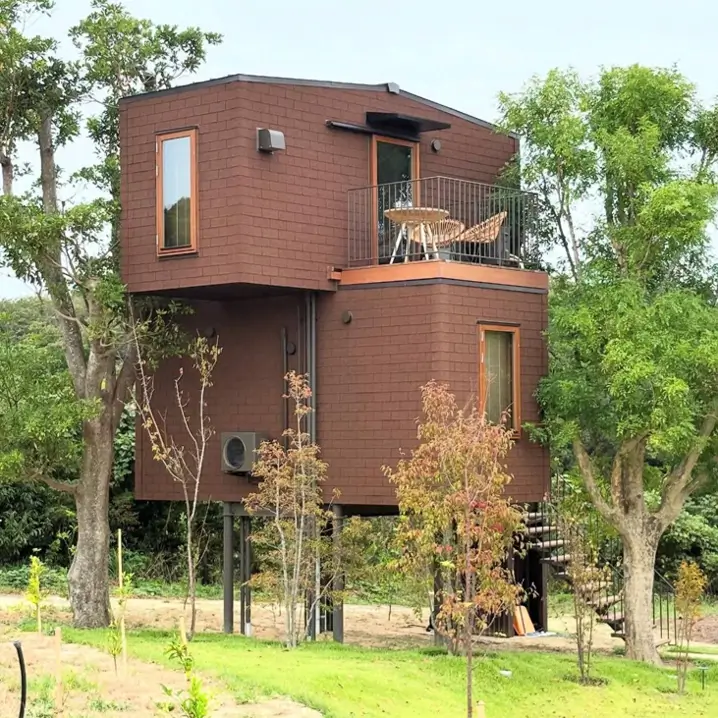 Modern brown shingled house on stilts surrounded by green trees with a small balcony and spiral staircase.