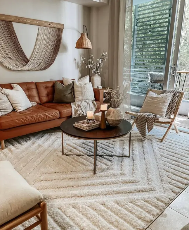 A warm neutral living room with a brown leather sofa, a high-pile geometric rug, and fiber wall art above the couch.