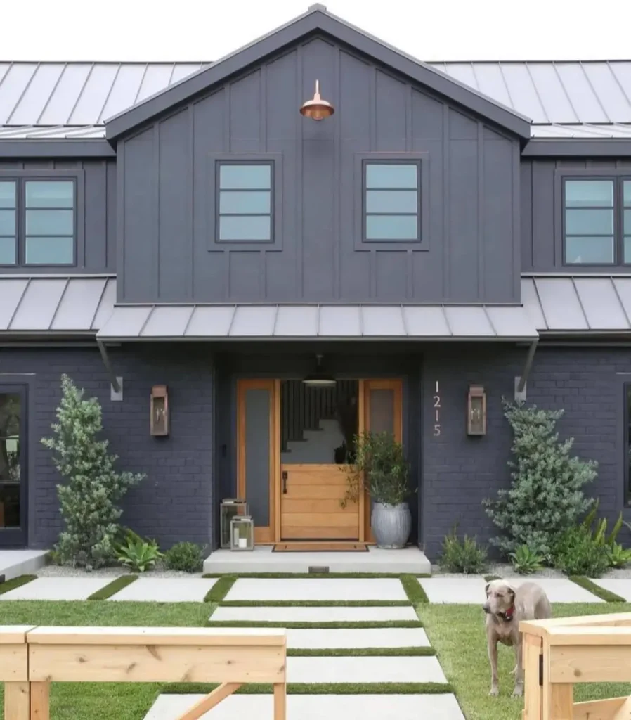 A modern black farmhouse with a light metal roof, a natural wood Dutch door, and a copper gooseneck light.