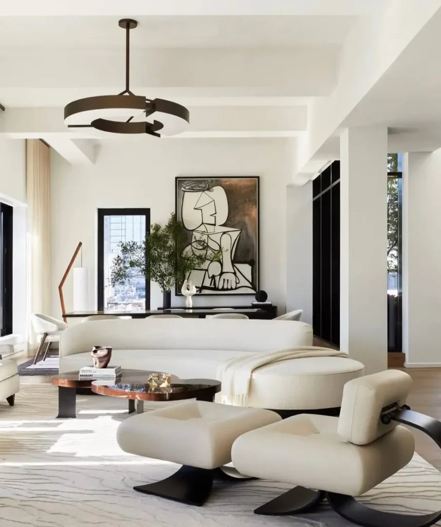 A modern, high-ceiling neutral living room with a large abstract painting, a curved white sofa, and sculptural armchairs.