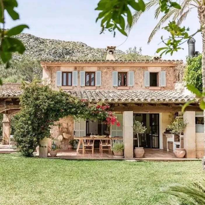 A rustic Mediterranean stone house with light blue shutters, a terracotta roof, and a covered patio.