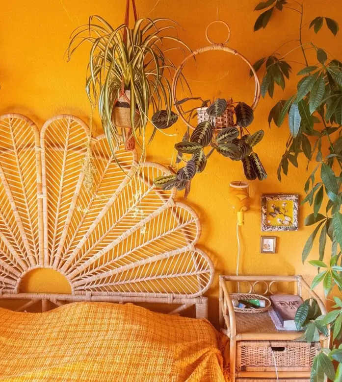 A bedroom with 70s interior decor, featuring an ornate rattan peacock headboard against a golden-yellow wall with houseplants.