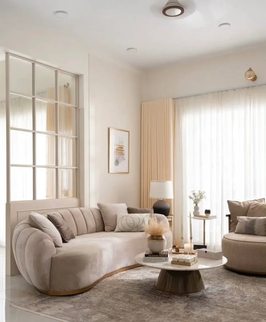 A warm neutral living room with a curved, channel-tufted beige sofa, a round marble coffee table, and an interior window wall.