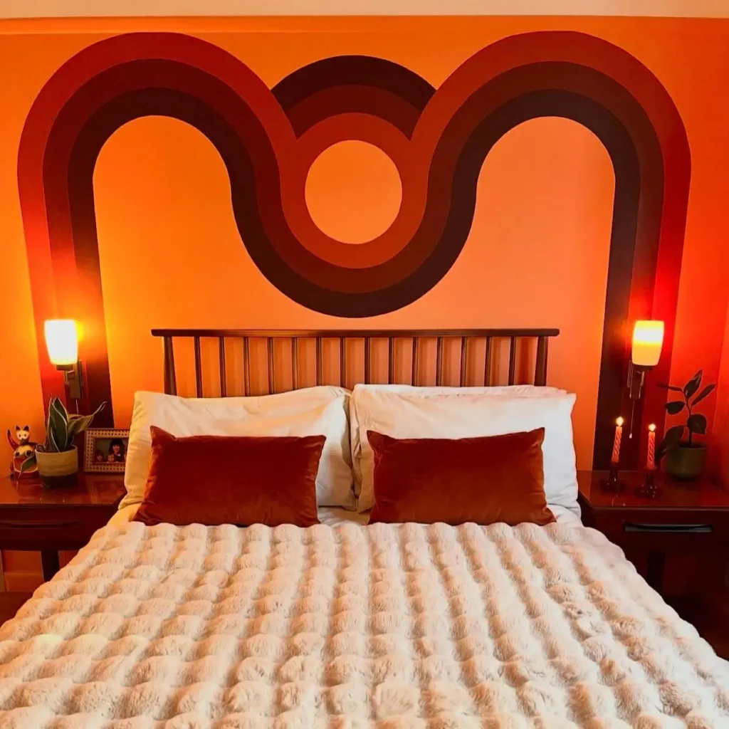 A 70s bedroom inspo wall with a painted graphic mural in orange and brown behind a bed.