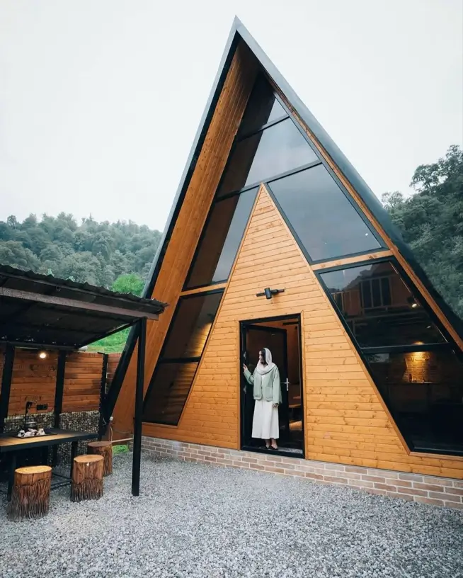 A modern minimalist A-frame house with a wood exterior and large triangular windows in a wooded setting.