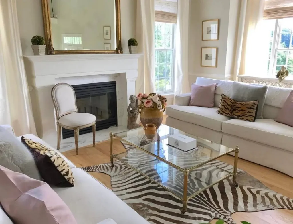 A classic neutral living room featuring white sofas, a zebra print rug, and a large gold mirror over the fireplace.