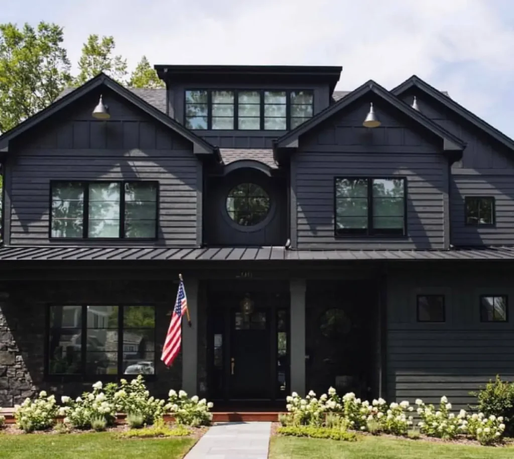 A symmetrical black farmhouse exterior with a central round window, horizontal siding, and a dark stone foundation.
