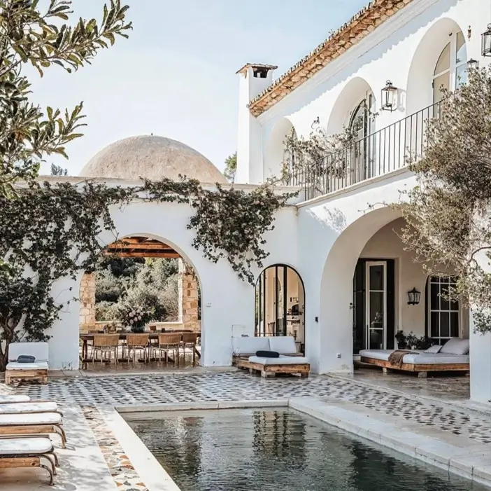 A Mediterranean pool courtyard with white stucco arches, patterned tile, and modern wooden daybeds.