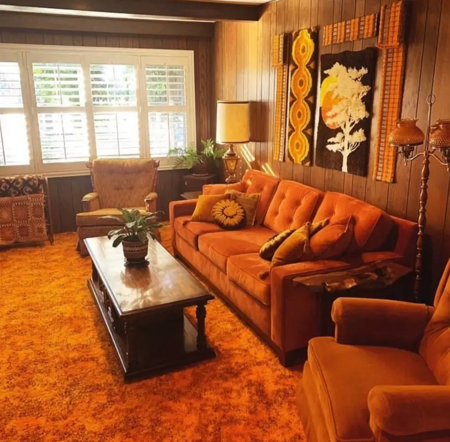A 70s living room with dark wood paneling, an orange sofa, and textile wall hangings.