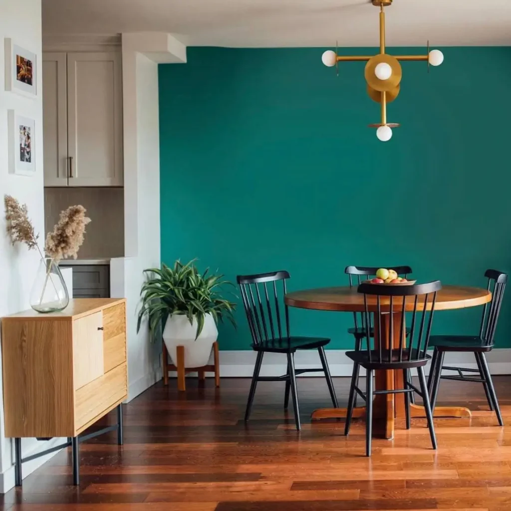 A round wood condo dining table with black chairs set against a bold teal accent wall in a modern dining room.