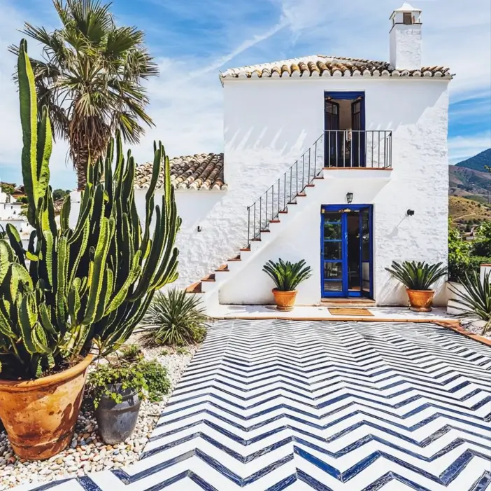A white Mediterranean house with a blue and white chevron tile patio, blue doors, and large potted cacti.