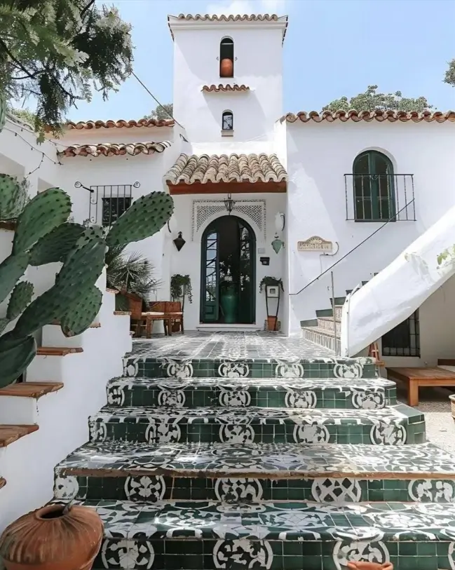 A white Mediterranean house with an elaborate green and white patterned tile staircase and dark green arched door.