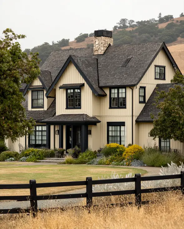 A modern beige farmhouse with black trim and a large stone chimney, set in a rural landscape.