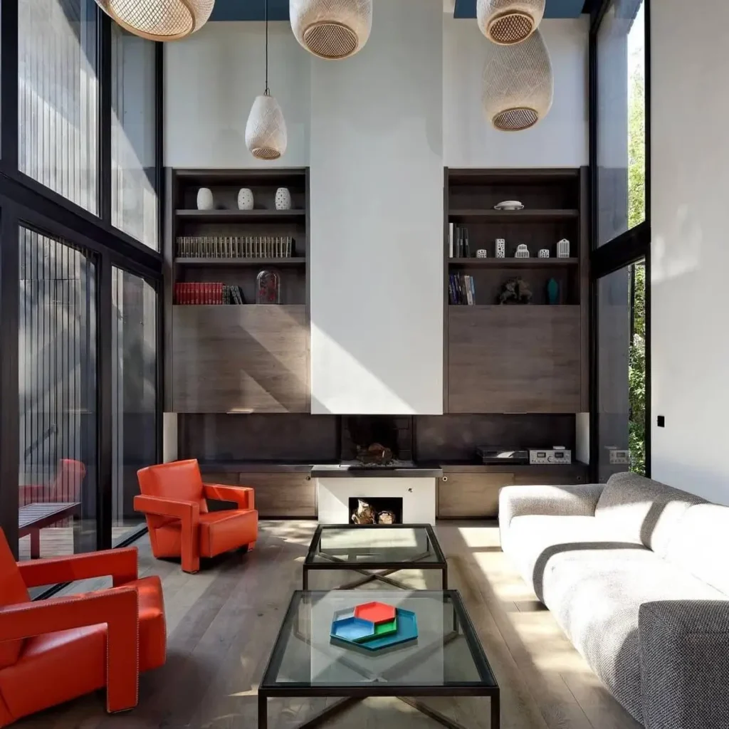 Minimalist living room with high ceilings, large windows, a central fireplace, and bright orange armchairs.