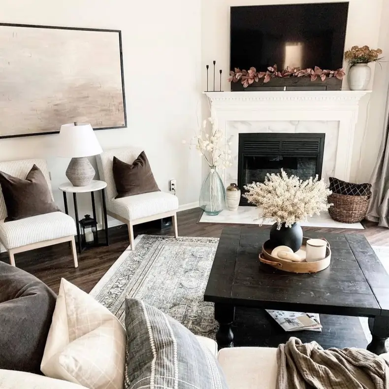 A cozy neutral living room centered around a classic white fireplace, with a dark wood coffee table and comfortable seating.